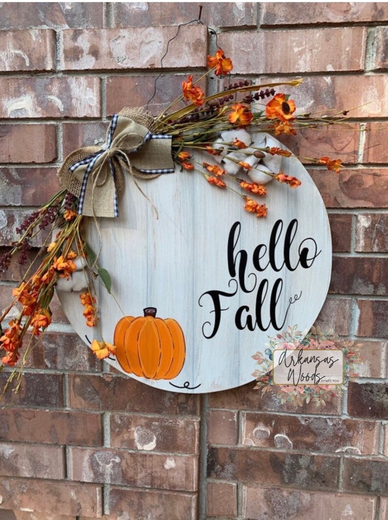 May include: A white wooden round door hanger with the text "hello fall" in black script. The door hanger is decorated with a burlap bow, orange and white flowers, and a painted orange pumpkin.