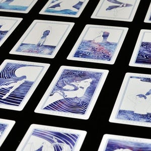 以下が含まれることがあります： 水彩イラストが描かれたタロットカードのセット。各カードには、水中の人物が描かれており、青と紫の色合いです。カードは黒い表面に配置されています。