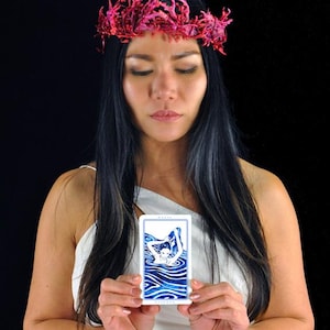 以下が含まれることがあります： ピンクの花の冠と白いトップスを着た女性が、水中の人物の青と白のイラストが描かれたタロットカードを持っています。背景は黒です。