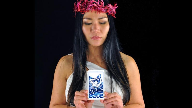 以下が含まれることがあります： ピンクの花の冠と白いトップスを着た女性が、水中の人物の青と白のイラストが描かれたタロットカードを持っています。背景は黒です。