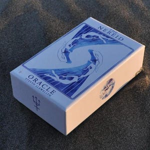 以下が含まれることがあります： 「The Nereid Oracle」のカードが入った白い箱。箱には、水中の人物の青いイラストと「Oracle」の文字が描かれています。箱は暗く、質感のある表面に置かれています。