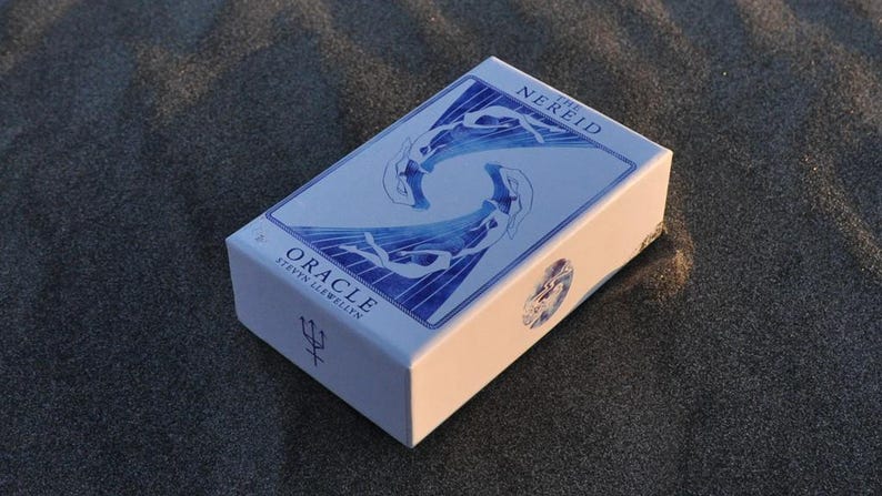 以下が含まれることがあります： 「The Nereid Oracle」のカードが入った白い箱。箱には、水中の人物の青いイラストと「Oracle」の文字が描かれています。箱は暗く、質感のある表面に置かれています。