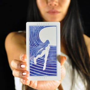 以下が含まれることがあります： 白い背景と青いイラストのタロットカード。カードには長い髪の女性と波のデザインが描かれています。「MENIPPE」と「FURY」の文字がカードに印刷されています。カードは黒い背景の前に保持されています。
