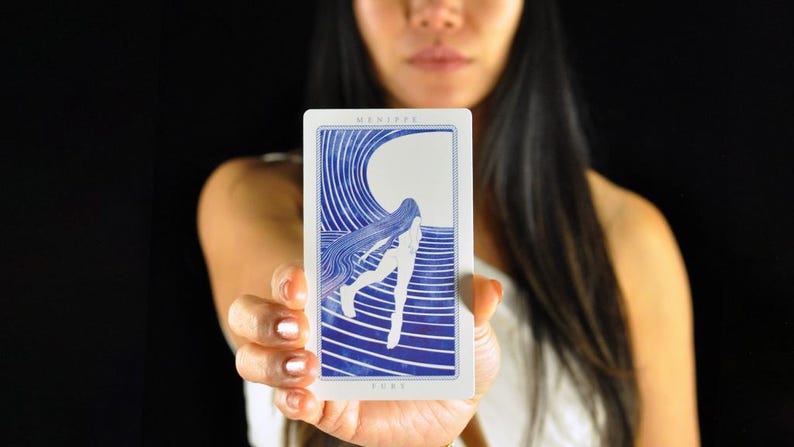 以下が含まれることがあります： 白い背景と青いイラストのタロットカード。カードには長い髪の女性と波のデザインが描かれています。「MENIPPE」と「FURY」の文字がカードに印刷されています。カードは黒い背景の前に保持されています。