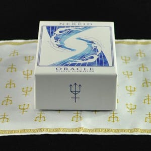 以下が含まれることがあります： 青と白のアートワークが施された「Nereid Oracle」カードの白い箱。箱は、金の三叉模様の白い布の上にあります。箱には三叉のシンボルがあります。