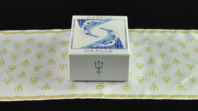 以下が含まれることがあります： 青と白のアートワークが施された「Nereid Oracle」カードの白い箱。箱は、金の三叉模様の白い布の上にあります。箱には三叉のシンボルがあります。