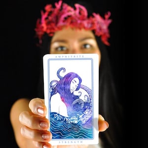 以下が含まれることがあります： 「AMPHITRITE」の文字が上部に、「STRENGTH」の文字が下部に書かれたタロットカード。紫色の髪と触手を持つ女性が、青と白の海の背景に描かれています。カードを持っている人。