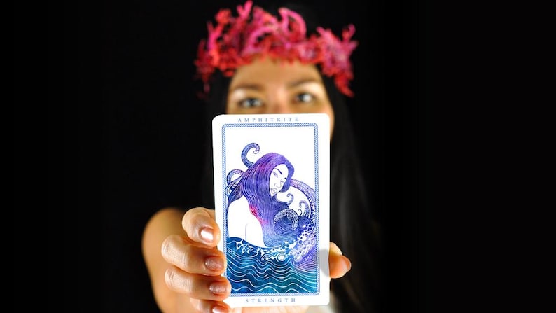 以下が含まれることがあります： 「AMPHITRITE」の文字が上部に、「STRENGTH」の文字が下部に書かれたタロットカード。紫色の髪と触手を持つ女性が、青と白の海の背景に描かれています。カードを持っている人。