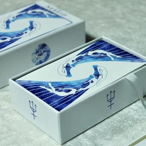 以下が含まれることがあります： 青と白のアートワークが施された2つの白いオラクルカードボックス。ボックスにはトライデントのシンボルと「Oracle」の文字が印刷されています。上部のボックスには「Nereid」の文字が印刷されています。ボックス内のカードは青と白のデザインです。