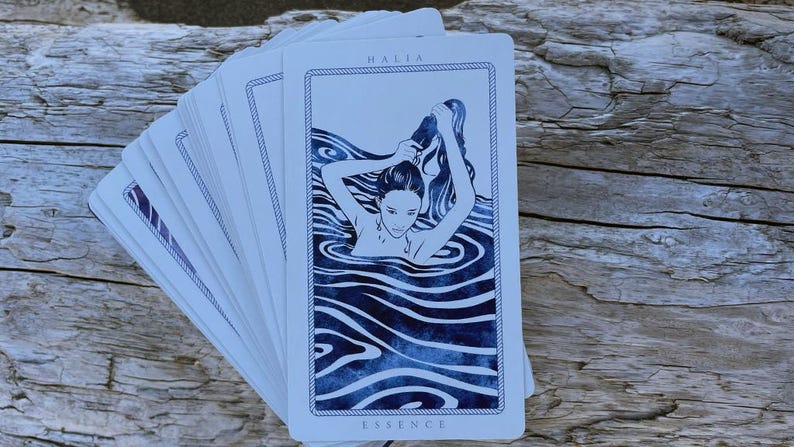 以下が含まれることがあります： 水中の女性の青と白のイラストが描かれたタロットカードの束。「HALIA」が上部に、「ESSENCE」が下部に記されています。カードは、風化した木製の表面に扇状に広げられています。
