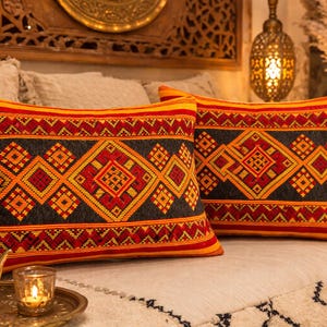 Puede incluir: Dos cojines decorativos rectangulares con un patrón geométrico en rojo, negro y naranja. Los cojines están sobre una superficie blanca. Los cojines tienen un diseño tradicional y forman parte de un tema de decoración del hogar.