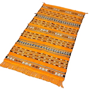 May include: A woven rug with a geometric pattern in shades of orange, brown, and white. The rug has fringe on all sides.