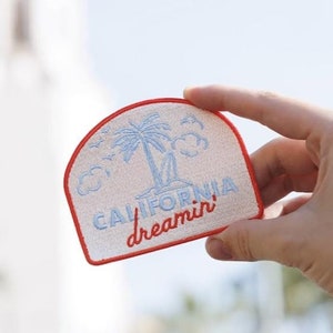May include: A white embroidered patch with red stitching. The patch features a palm tree, clouds, and the text "California dreamin'" in blue lettering.