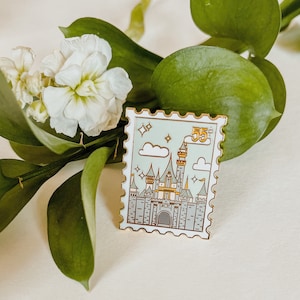May include: A gold-colored enamel pin shaped like a postage stamp with a cartoon illustration of a castle with a green and white color scheme. The stamp has the text "55¢" in gold lettering.