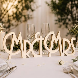 May include: Wooden "Mr & Mrs" sign in a cursive font, placed on a white tablecloth. Two champagne flutes stand behind the sign. The table is set with white plates, silverware, and floral arrangements, creating a romantic wedding theme.