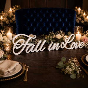 Peut inclure: Une enseigne en bois blanc avec le texte cursif Fall in Love, posée sur une table sombre. L'enseigne est entourée de fleurs blanches, de feuillage vert et de bougies allumées dans des photophores, avec une chaise bleue capitonnée en arrière-plan.