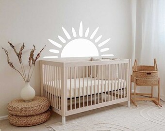 May include: A white cot with a white mattress and a white sun decal on the wall behind it. The cot is in a nursery with a white rug, a wicker basket, and a beige vase with dried flowers.