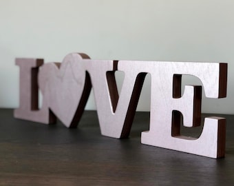 May include: Wooden letters spelling "LOVE" in a light pink color with a heart replacing the "O". The letters are on a dark wood surface.