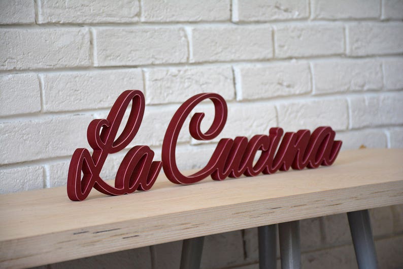 Puede incluir: Un letrero de madera rojo con las palabras "La Cucina" en una fuente cursiva. El letrero est&aacute; sobre una mesa de madera clara.