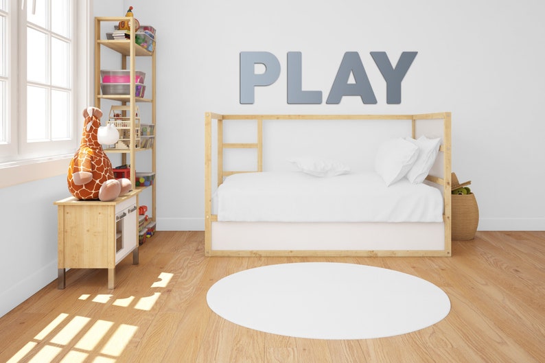 May include: A white bed with a wooden frame and a white rug in front of it. The wall behind the bed has the word "PLAY" written in large, gray letters.