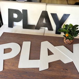 May include: Two sets of the word "PLAY" spelled out in large, block letters. One set is black and the other is white. The white letters are on a wooden table, and the black letters are on a white couch.