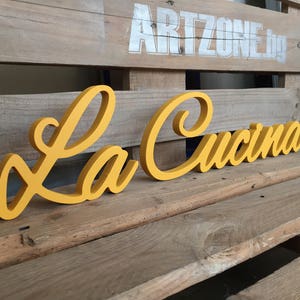 Puede incluir: Un letrero de madera amarillo con las palabras "La Cucina" en una fuente cursiva.
