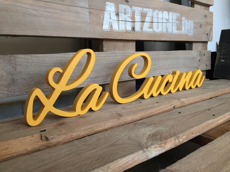 Puede incluir: Un letrero de madera amarillo con las palabras "La Cucina" en una fuente cursiva.