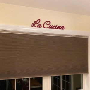 Puede incluir: Un letrero de madera rojo con las palabras "La Cucina" en escritura cursiva. El letrero est&aacute; montado en una pared blanca sobre una persiana de ventana marr&oacute;n.