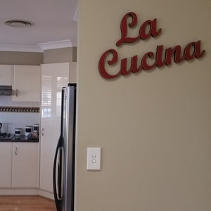 Puede incluir: Una cocina blanca con una nevera de acero inoxidable y un letrero que dice "La Cucina" en letras rojas.