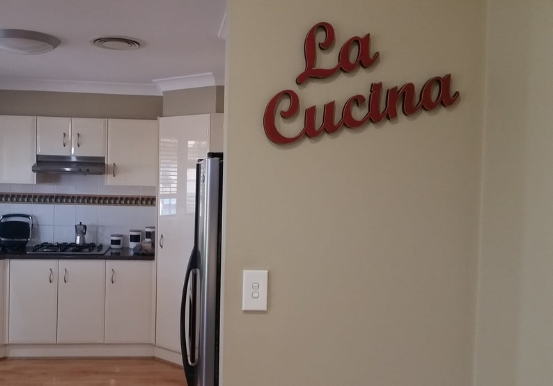 Puede incluir: Una cocina blanca con una nevera de acero inoxidable y un letrero que dice "La Cucina" en letras rojas.