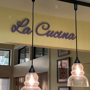 Puede incluir: Tres l&aacute;mparas colgantes de vidrio transparente con una superficie texturizada cuelgan de cables negros contra una pared amarilla. El letrero "La Cucina" est&aacute; escrito en letras cursivas moradas sobre las luces.