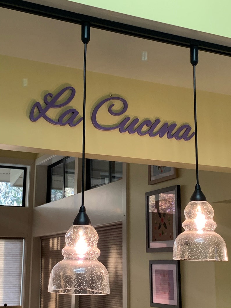 Puede incluir: Tres l&aacute;mparas colgantes de vidrio transparente con una superficie texturizada cuelgan de cables negros contra una pared amarilla. El letrero "La Cucina" est&aacute; escrito en letras cursivas moradas sobre las luces.