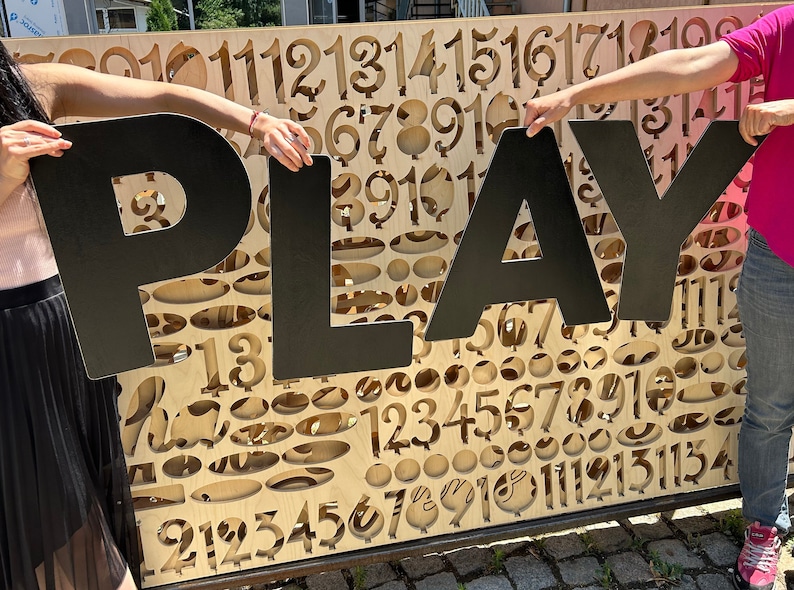 May include: Two people holding up large black letters that spell out the word "PLAY" in front of a wooden wall with cut-out numbers.