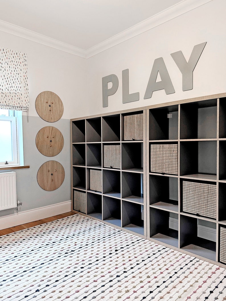May include: A playroom with a white and gray color scheme. The room features a large bookshelf with gray shelves and woven baskets. The wall is decorated with the word "PLAY" in large gray letters and three wooden circles with a textured surface. A white rug with black and pink dots covers the floor.