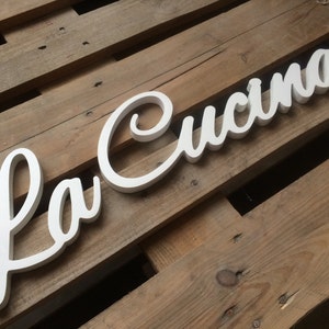 Puede incluir: Un letrero de madera blanco con las palabras "La Cucina" en una fuente cursiva.