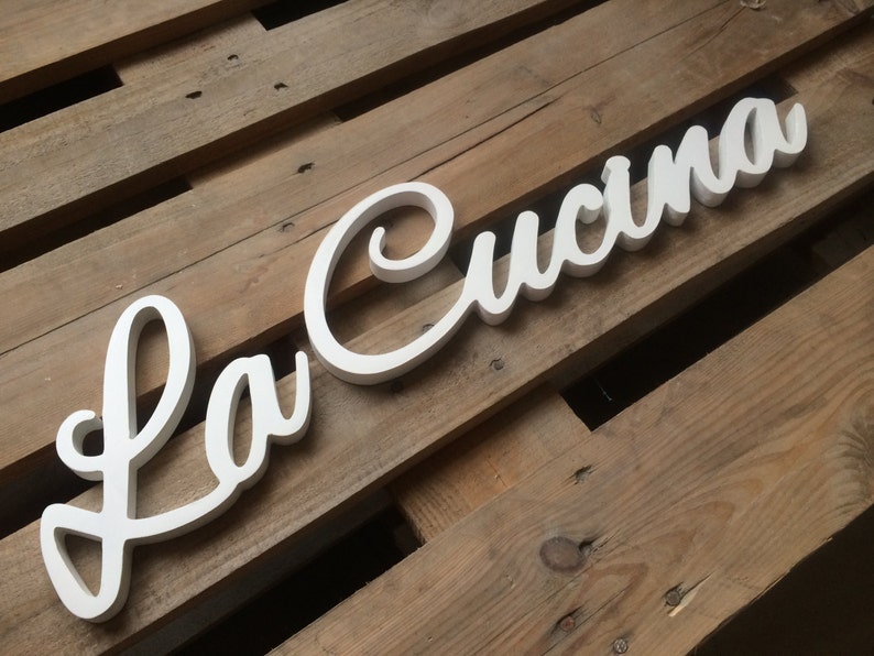Puede incluir: Un letrero de madera blanco con las palabras "La Cucina" en una fuente cursiva.