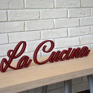 Puede incluir: Un letrero de madera rojo con las palabras "La Cucina" en una fuente cursiva.