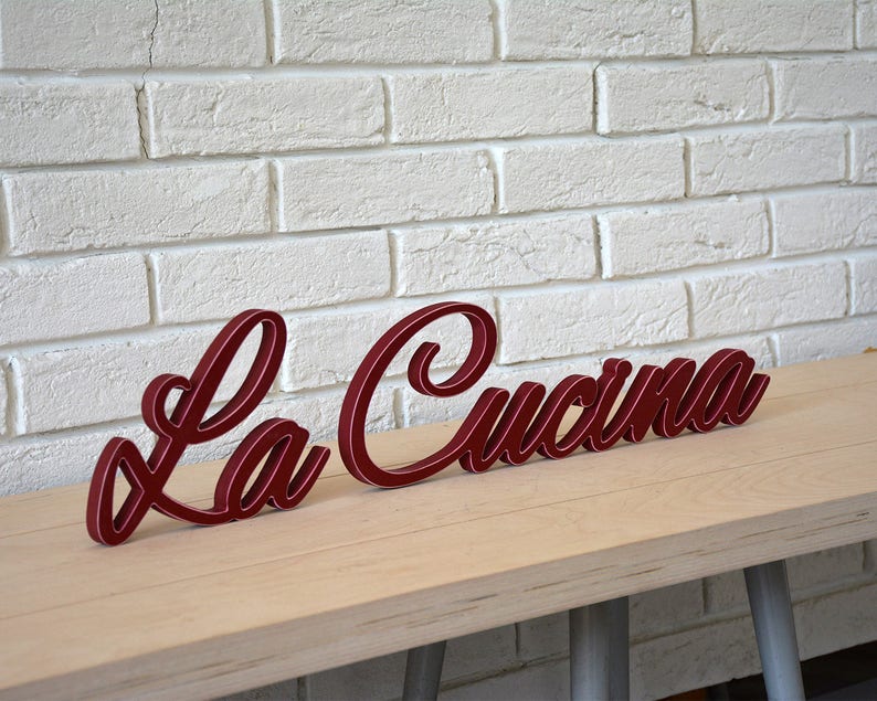 Puede incluir: Un letrero de madera rojo con las palabras "La Cucina" en una fuente cursiva.