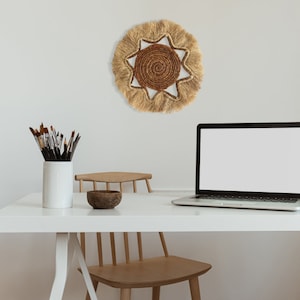 May include: A white desk with a laptop, a bowl, and a set of paintbrushes in a white vase. A wooden chair with a woven sun-shaped wall hanging above it.