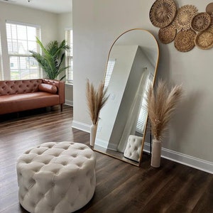 May include: A white tufted ottoman sits in front of a gold arched mirror in a room with light brown hardwood floors.  Dried pampas grass is in vases on either side of the mirror.  A brown leather sofa is visible in the background.
