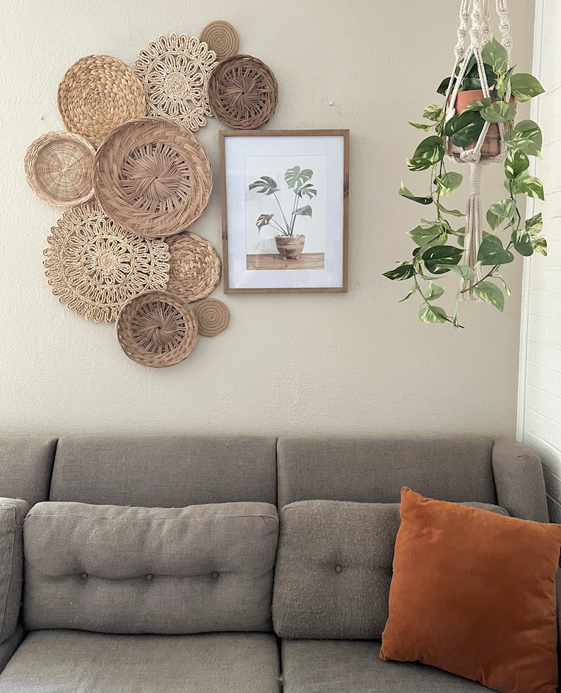 May include: A grey sofa with two cushions, one brown and one grey, in front of a wall with a framed print of a plant and a hanging macrame plant holder with a potted plant. The wall is decorated with a collection of woven wall decor.