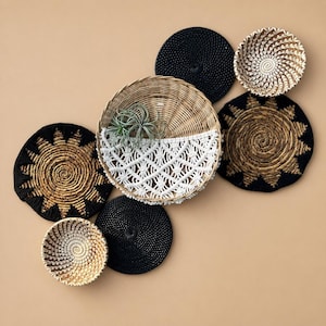 May include: A collection of wall decor items including two black circular woven discs, two small woven bowls with natural and white patterns, two sun-shaped woven wall hangings in black and gold, and a large central basket with macrame.