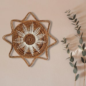 May include: A woven star-shaped wall hanging with a white sunburst design in the center. The wall hanging is made of natural materials and has a rustic, bohemian aesthetic.