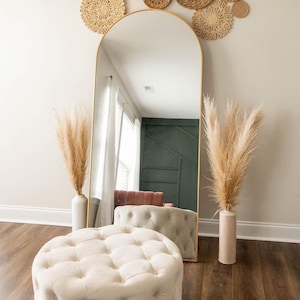 May include: A white tufted ottoman sits in front of a gold arched mirror with a wall decor of woven round baskets in shades of brown and beige.