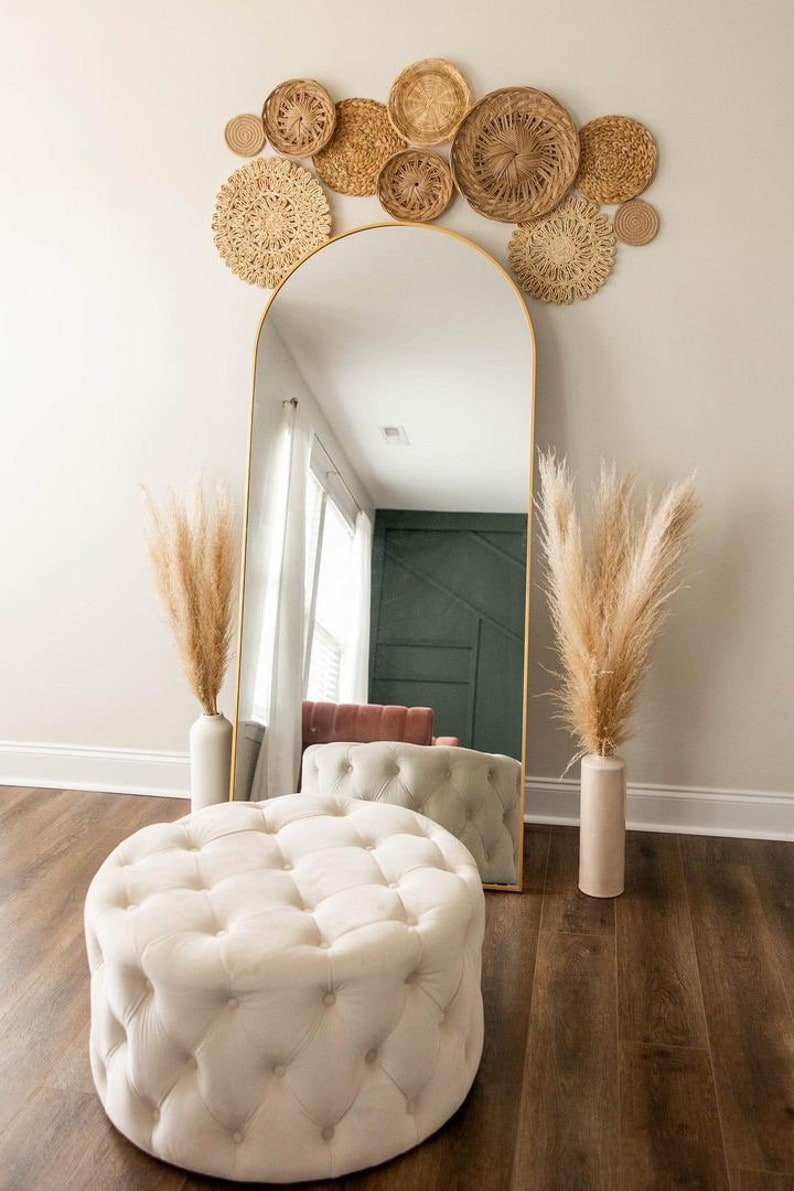 May include: A white tufted ottoman sits in front of a gold arched mirror with a wall decor of woven round baskets in shades of brown and beige.