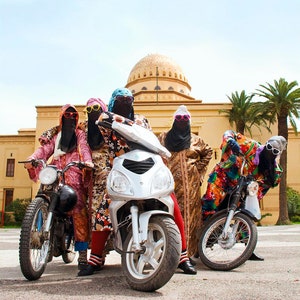 May include: Five people wearing colorful clothing and sunglasses are riding motorcycles in front of a building. The motorcycles are a silver scooter, a black motorcycle, and a red motorcycle.