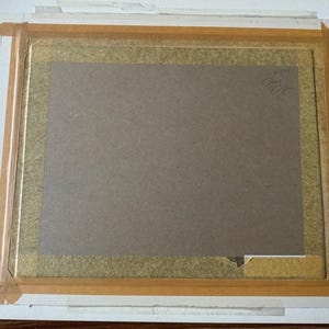 Puede incluir: La parte posterior de una obra de arte rectangular enmarcada con un soporte de cart&oacute;n marr&oacute;n asegurado con cinta adhesiva de color canela en los bordes. Una firma manuscrita y una peque&ntilde;a marca num&eacute;rica son visibles en la esquina superior.