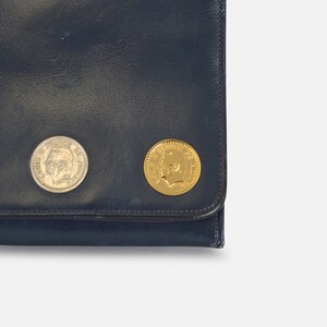 Puede incluir: Cartera de cuero azul oscuro con dos monedas adjuntas. Una moneda es plateada y la otra dorada. Ambas monedas presentan el perfil de una persona.