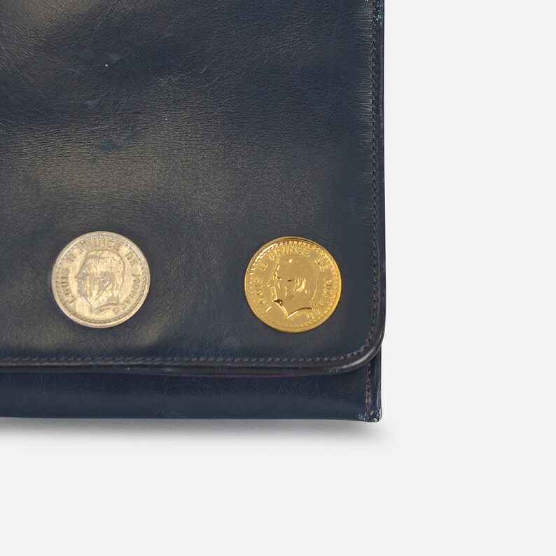 Puede incluir: Cartera de cuero azul oscuro con dos monedas adjuntas. Una moneda es plateada y la otra dorada. Ambas monedas presentan el perfil de una persona.