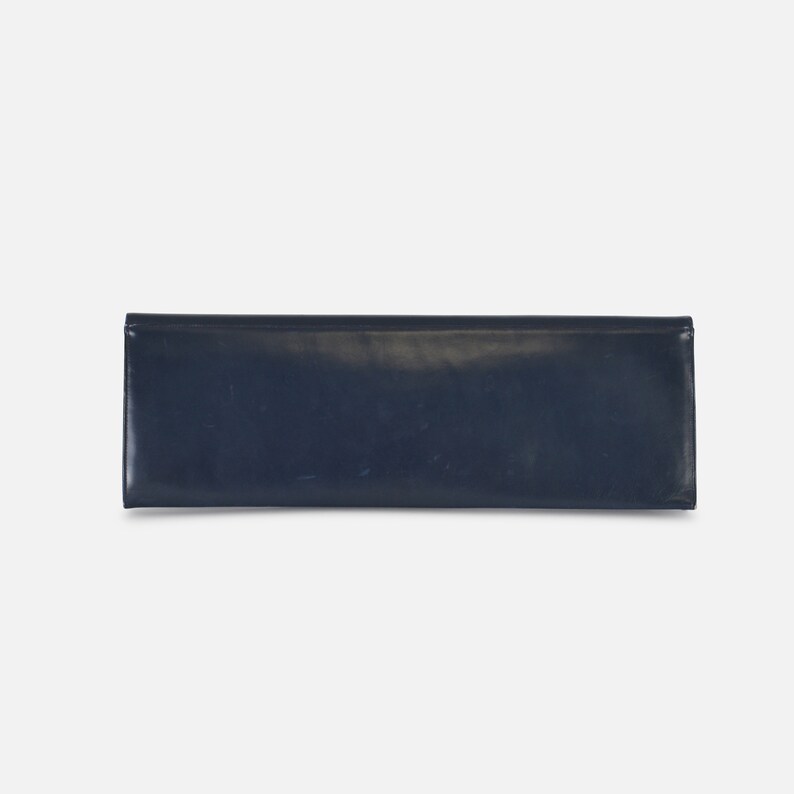 Puede incluir: Un bolso de mano azul marino de forma rectangular. El bolso tiene un acabado brillante y un dise&ntilde;o sencillo y elegante. El bolso de mano est&aacute; cerrado y parece vac&iacute;o.